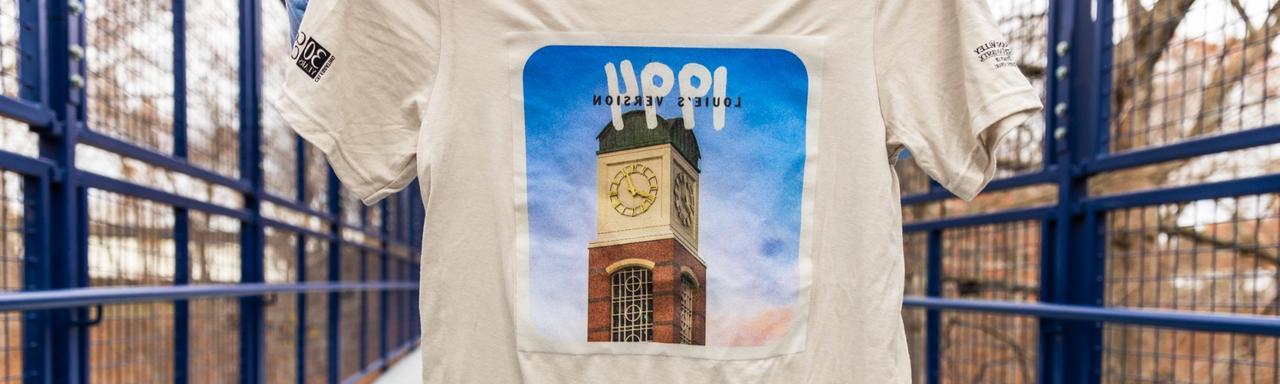 A student holding a beige t-shirt featuring a clock tower and text that reads "1994. Louie's version."